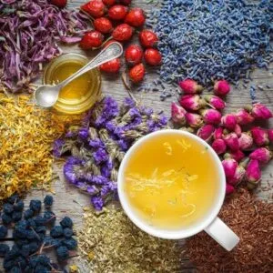 Foto dall'alto di un tavolo cosparso da erbe officinali in polvere sul quale sono poggiate una Tazza di tisana alla camomilla e un barattolino con un prodotto chiaro all'interno
